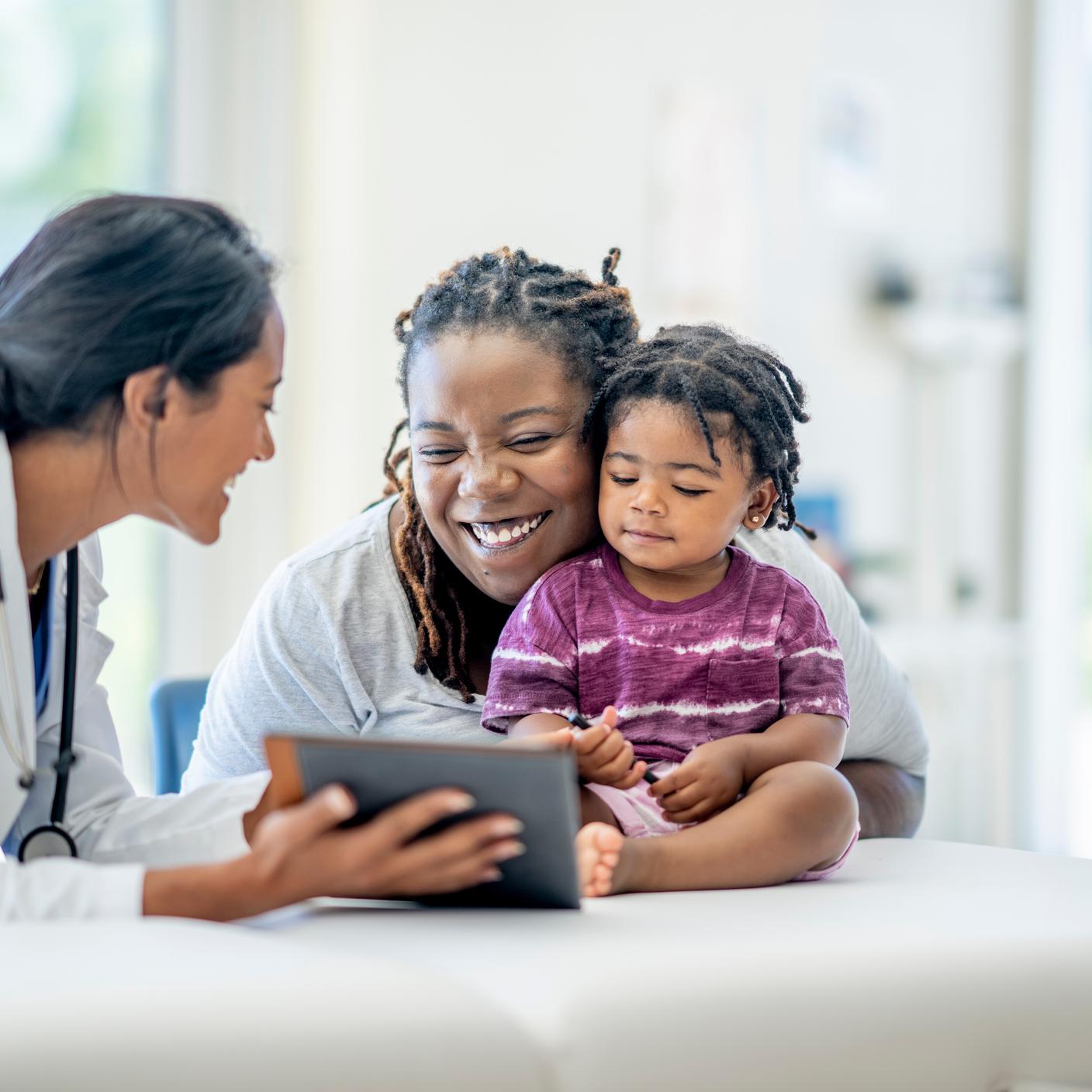 Một bà mẹ trẻ đưa con trai đi khám bác sĩ. Bé trai ngồi trên bàn khám bệnh, mẹ ngồi chắn đằng sau để đảm bảo bé không bị ngã. Nữ bác sĩ gốc Trung Đông ngồi trước mặt hai mẹ con, vừa mỉm cười vừa trò chuyện.