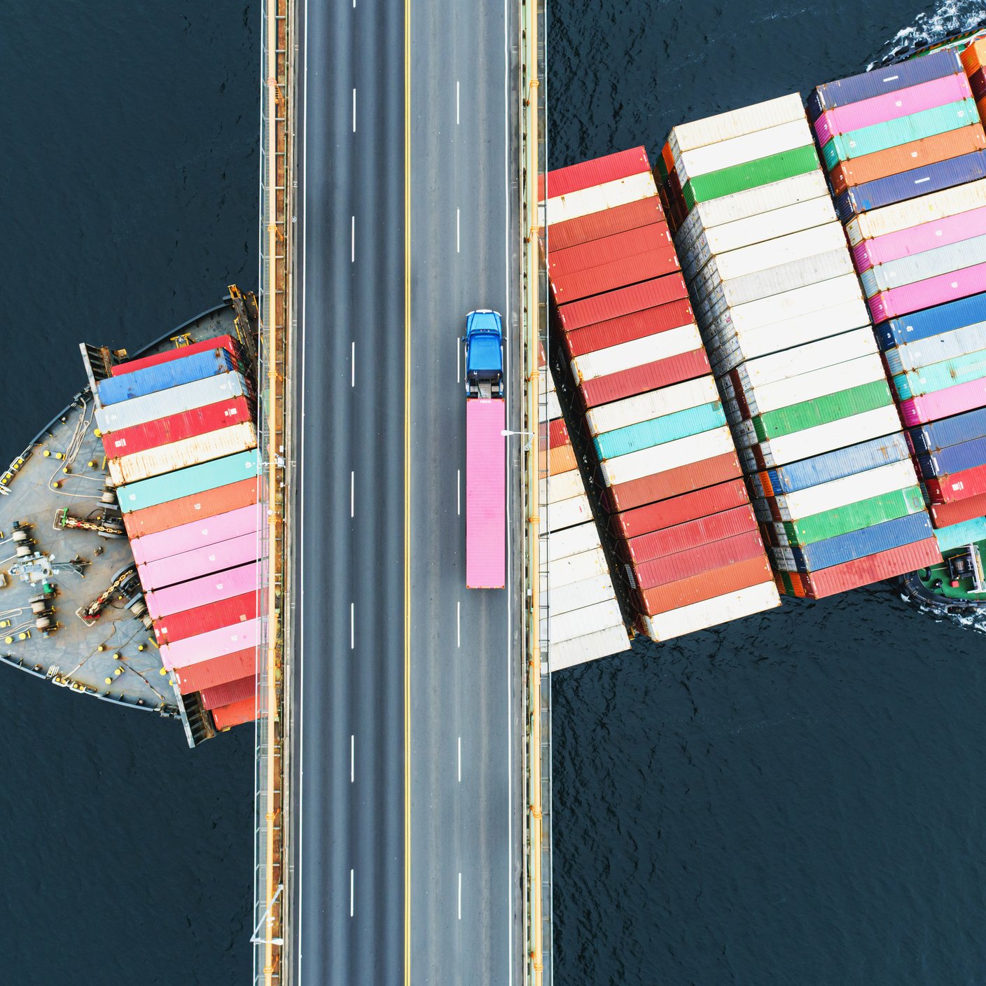 Aerial view of a container ship passing beneath a suspension bridge. Semi truck with pink cargo container crosses above.