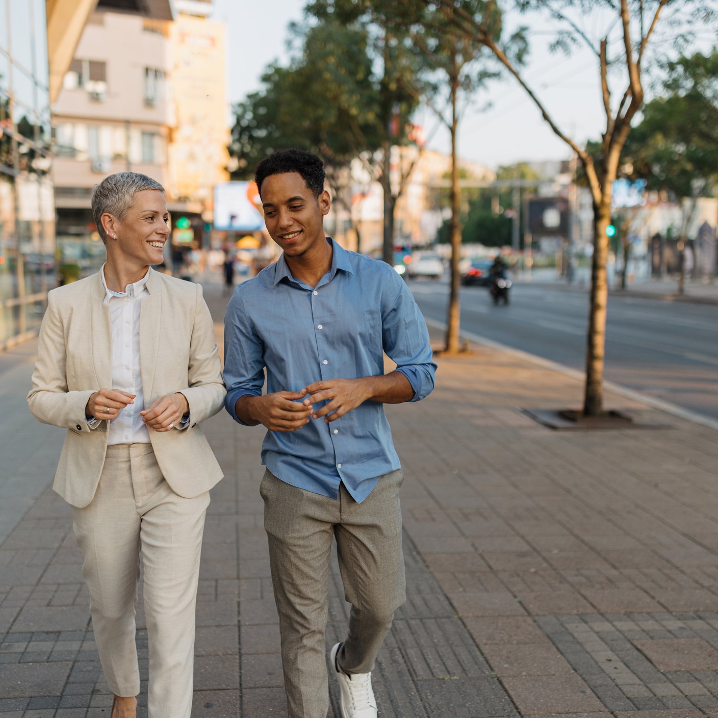 A businesswoman in a suit and a young man in casual clothing walking side by side in a city sidewalk, discussing with modern buildings and some vehicles in the background.