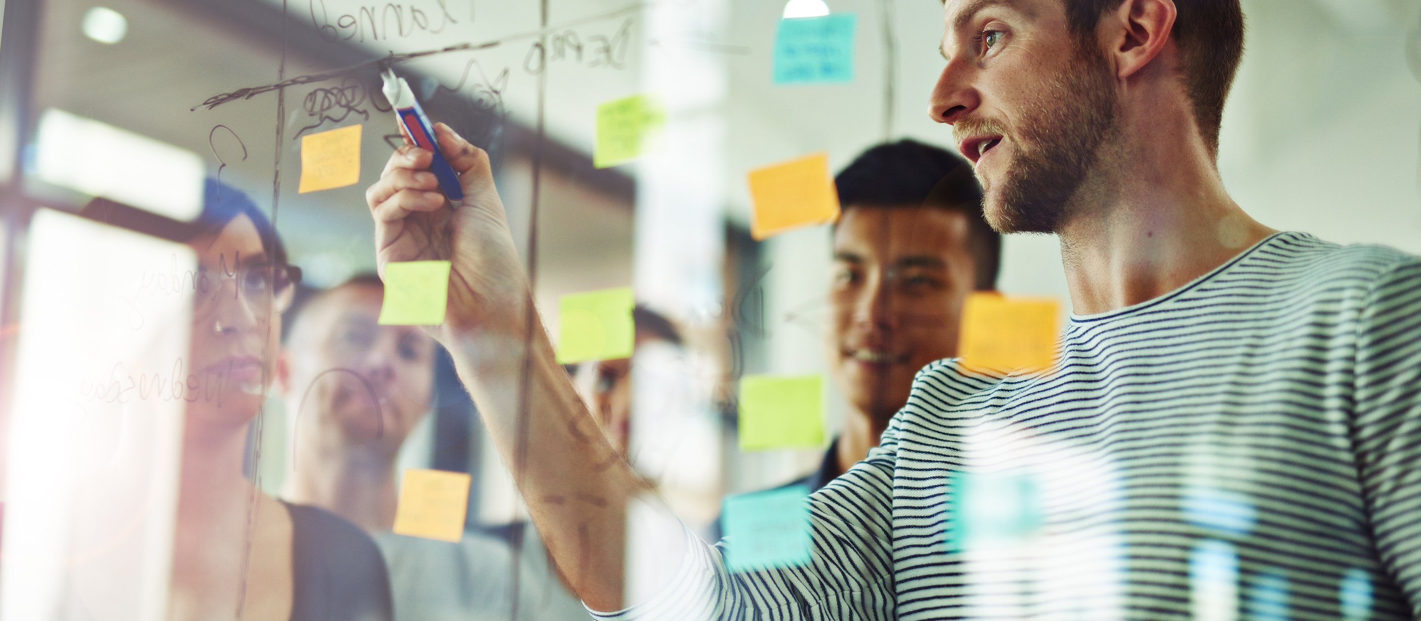 A person writes on a glass board with sticky notes while colleagues observe.