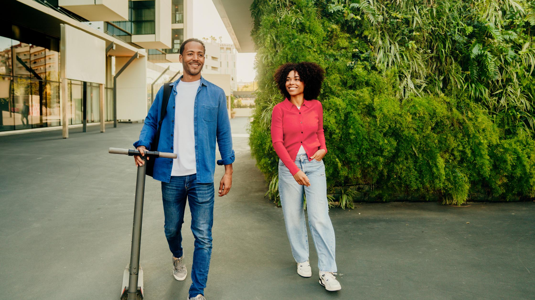 Couple smiling and walking in a green city space. The man is holding an e-scooter.